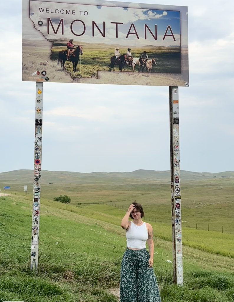 Chloë, wearing floral pants and a white tank top, stands before a "Welcome to Montana" sign and a green hillside. 
